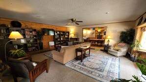 A cozy living room with a sofa, armchairs, a coffee table, and a bookshelf. There is a dining area with a table and chairs, and a kitchen in the background. - Inn the Clouds Hostel & Inn (Leadville)