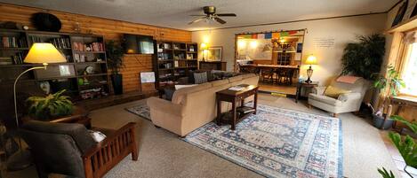 A cozy living room with a sofa, armchairs, a coffee table, and a bookshelf. There is a dining area with a table and chairs, and a kitchen in the background.
