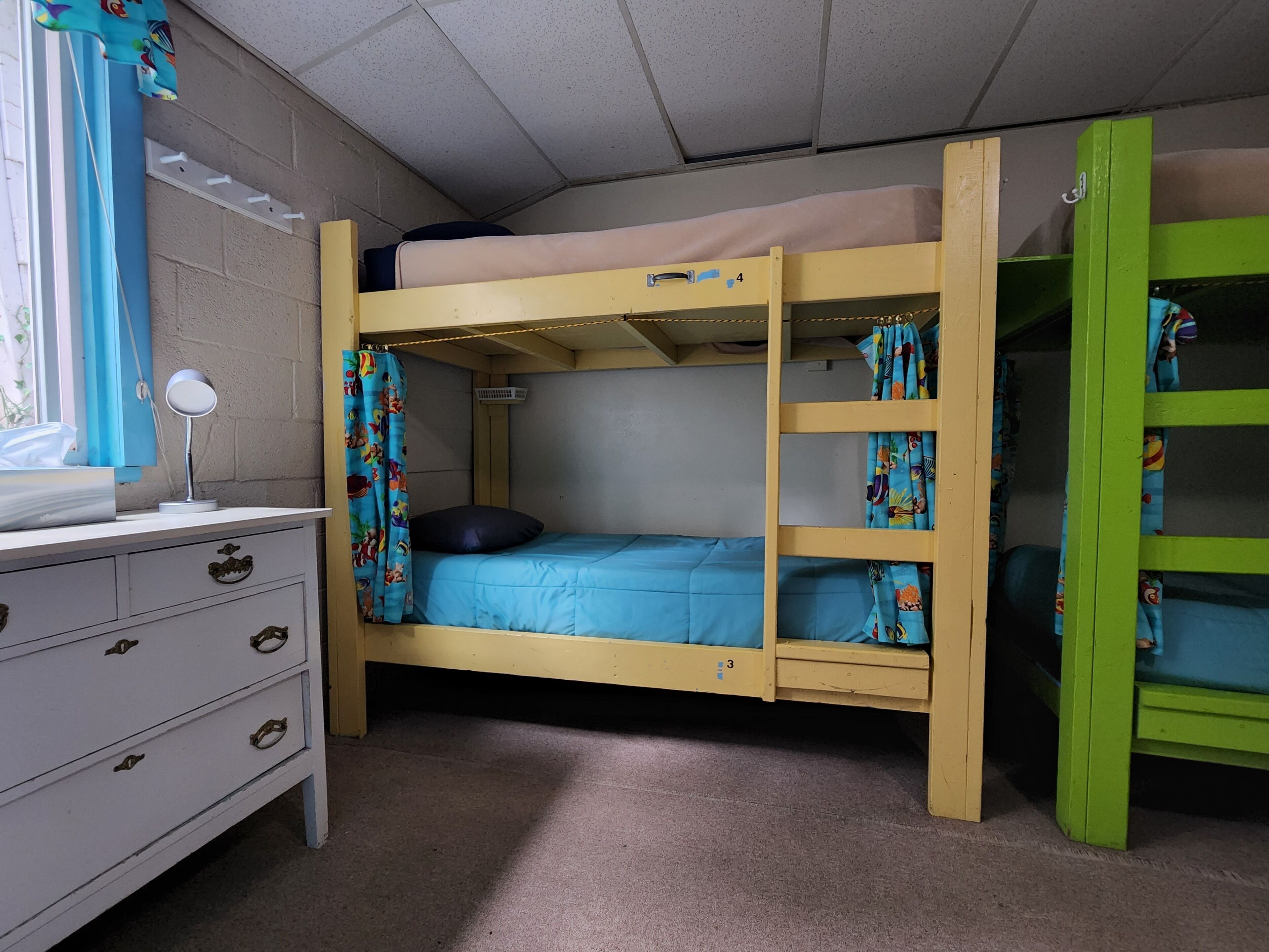 A bunk bed room with a desk and drawers, a window with curtains, and a green wardrobe.