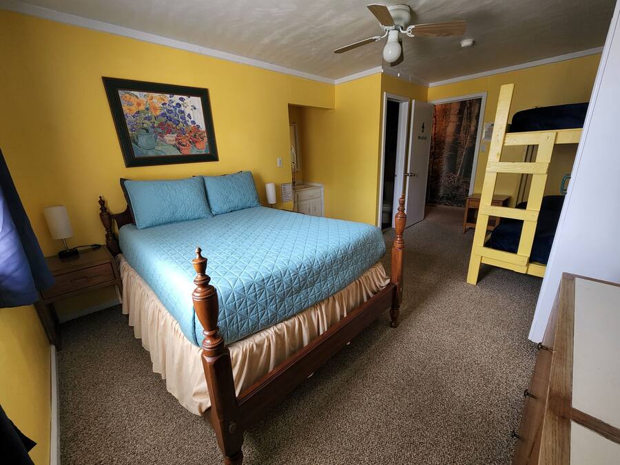Un dormitorio con cama de madera, ventilador de techo, un cuadro en la pared y una puerta que da a otra habitación.