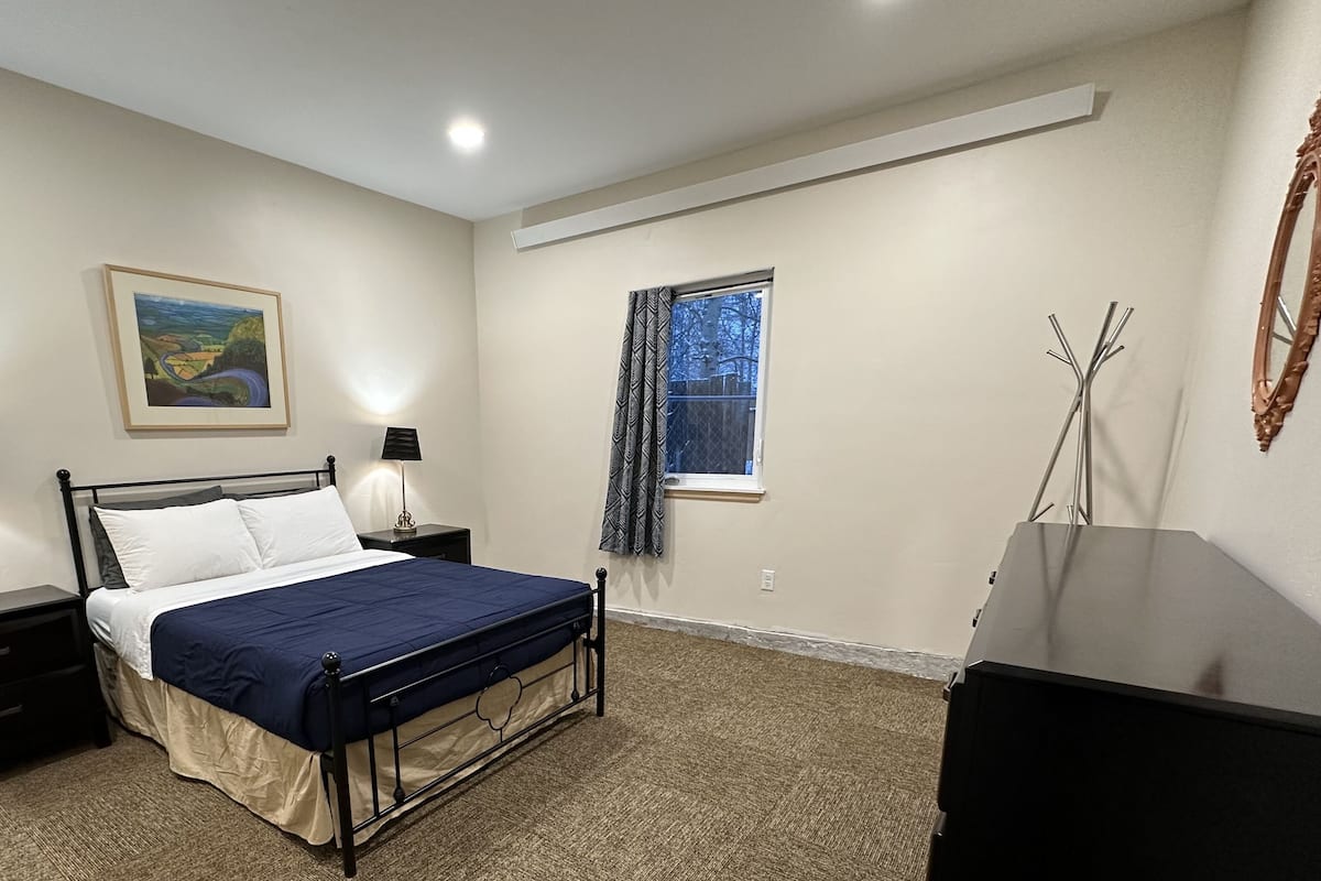 A bedroom with a bed, two bedside tables with lamps, a window with curtains, a painting on the wall, and a mirror on the right.
