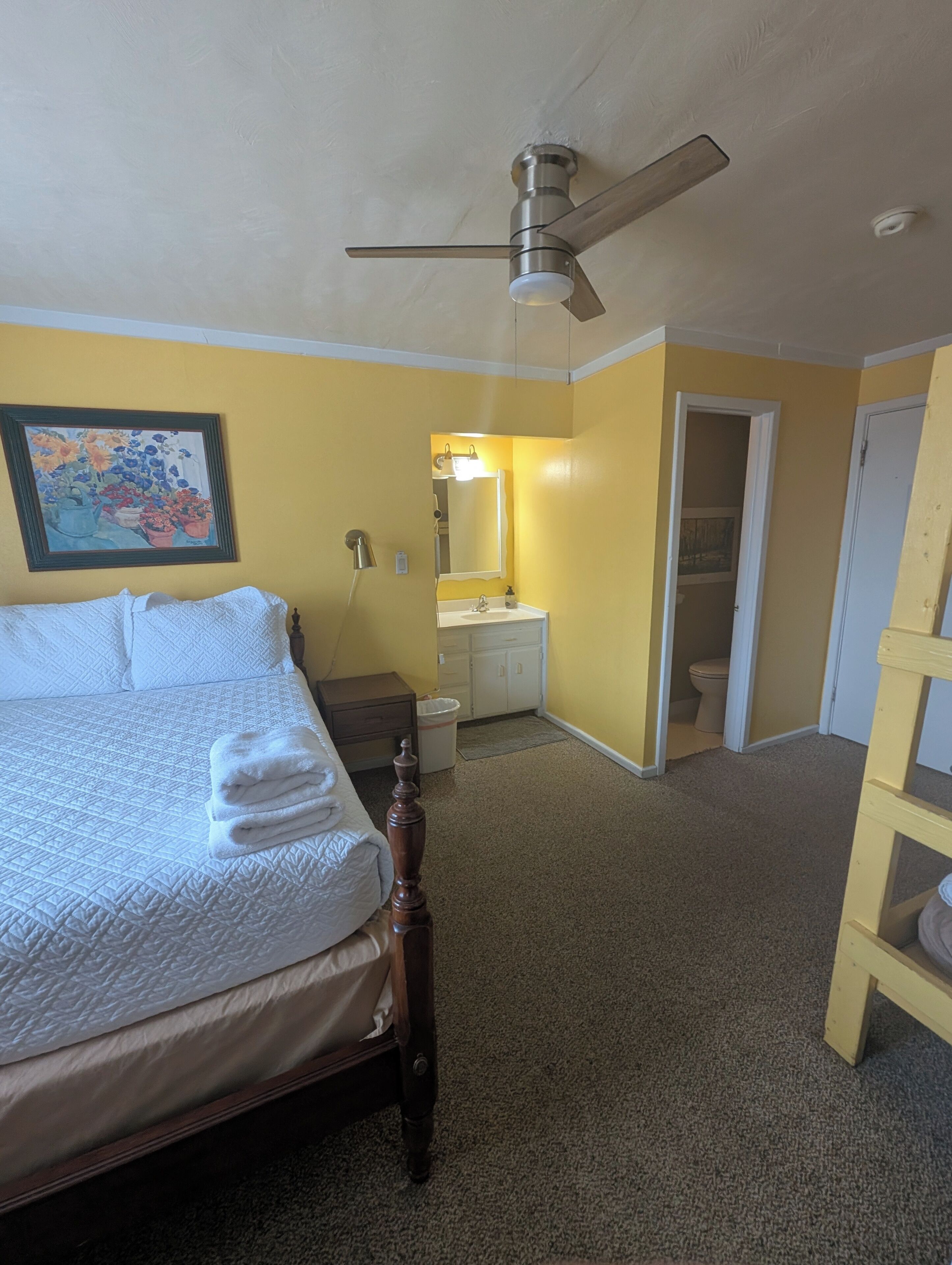 A bedroom with a bed, a ceiling fan, a bathroom with a sink and mirror, and a door leading to another room.
