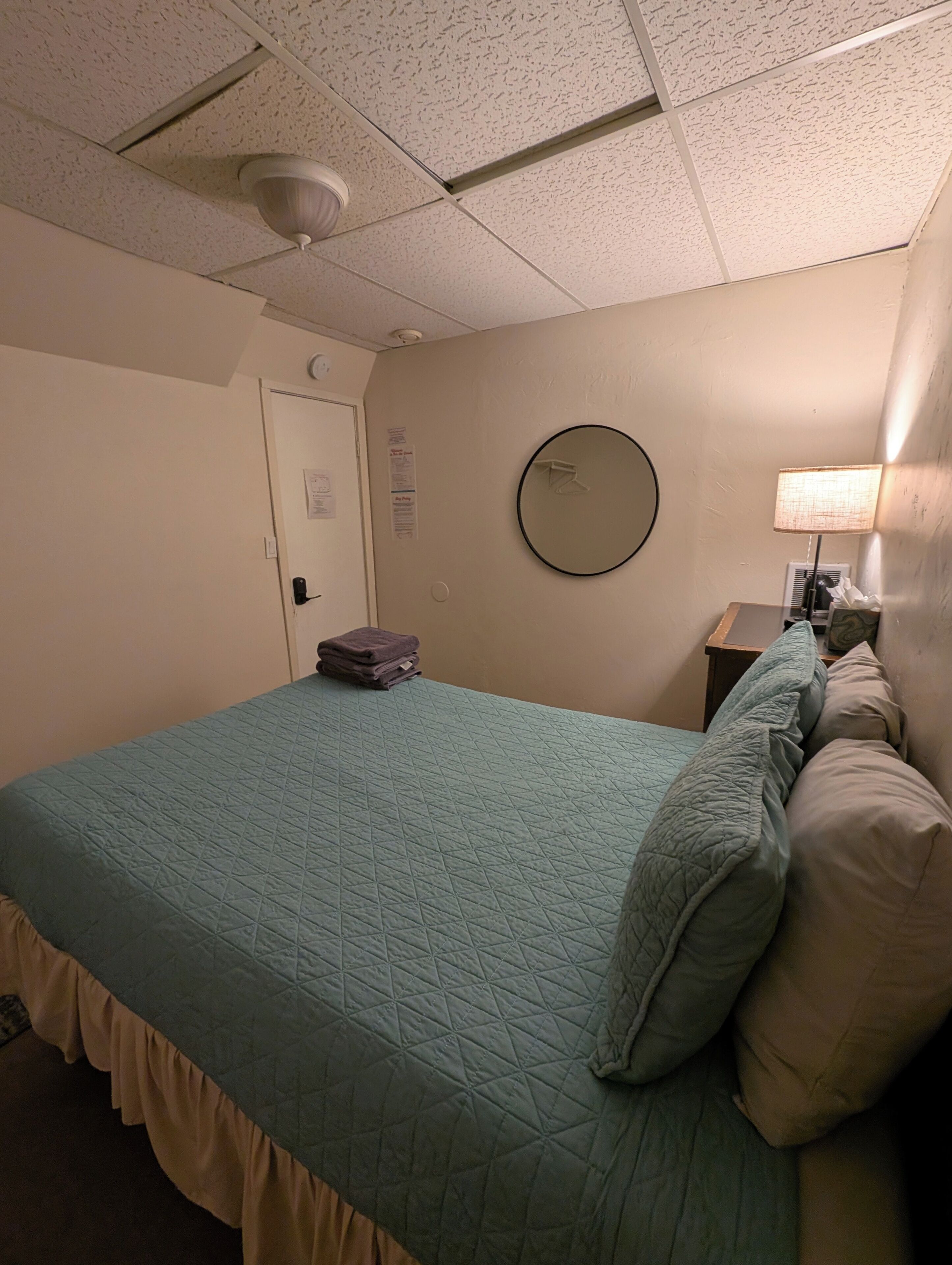 Una habitación de hotel con una cama, un espejo redondo, una lámpara y una puerta.