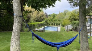 Una hamaca atada entre dos árboles en un patio trasero con una piscina y una terraza de madera al fondo.