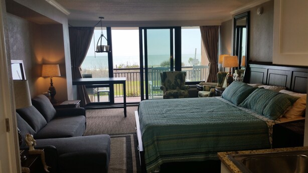 Ein Hotelzimmer mit einem großen Bett, einer Couch, einem kleinen Tisch, einem Sessel und Meerblick vom Balkon.