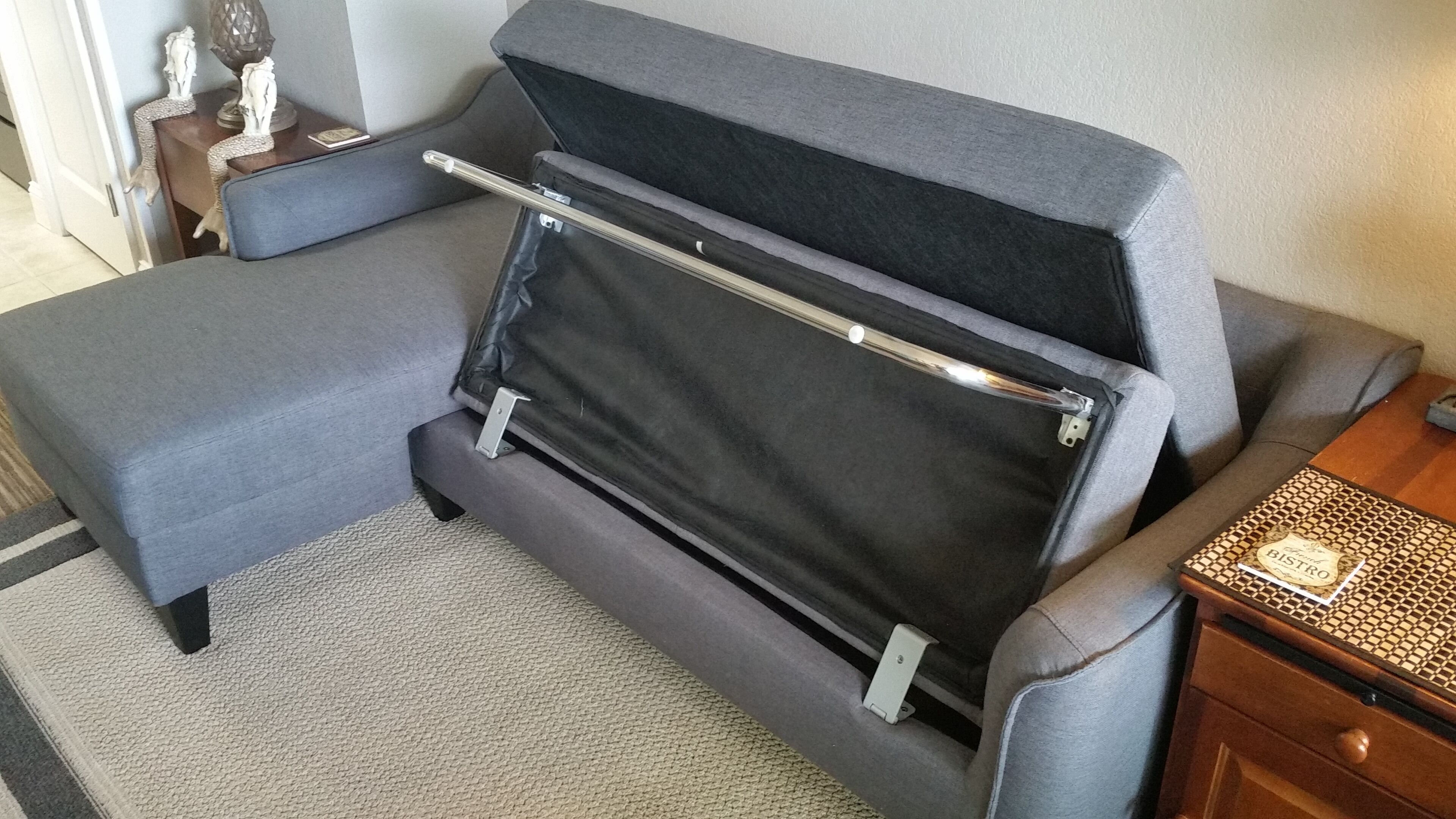 A compact, gray sofa with a pull-out table, a small wooden side table with a decorative item, and a patterned rug.