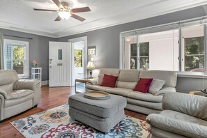 A living room with a sofa, ottoman, and armchair, a ceiling fan, and a door leading outside. - Welcome Home (+ your pup) MSU 2 mi (Lansing)