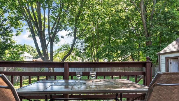 An outdoor patio with a wooden table and chairs, two wine glasses, and a view of a backyard with trees. - Welcome Home (+ your pup) MSU 2 mi (Lansing)