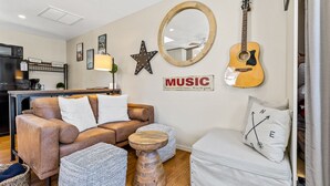Un salon chaleureux avec un canapé en cuir marron, une table basse en bois et un fauteuil blanc. Une guitare et une décoration en forme d’étoile sont accrochées au mur.