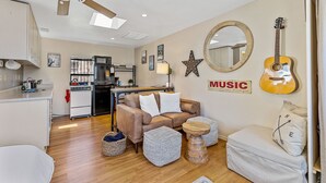 Un salon chaleureux avec un canapé, des poufs, une table basse, une guitare et, au fond, une cuisine.
