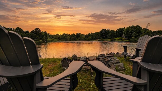 Adirondack-stole mod en sø med udsigt til solnedgang, omgivet af grønt og en stenvæg.