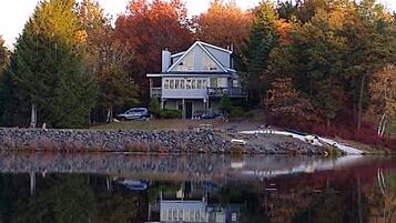 A lakeside house with a reflection in the water, surrounded by autumn-colored trees.