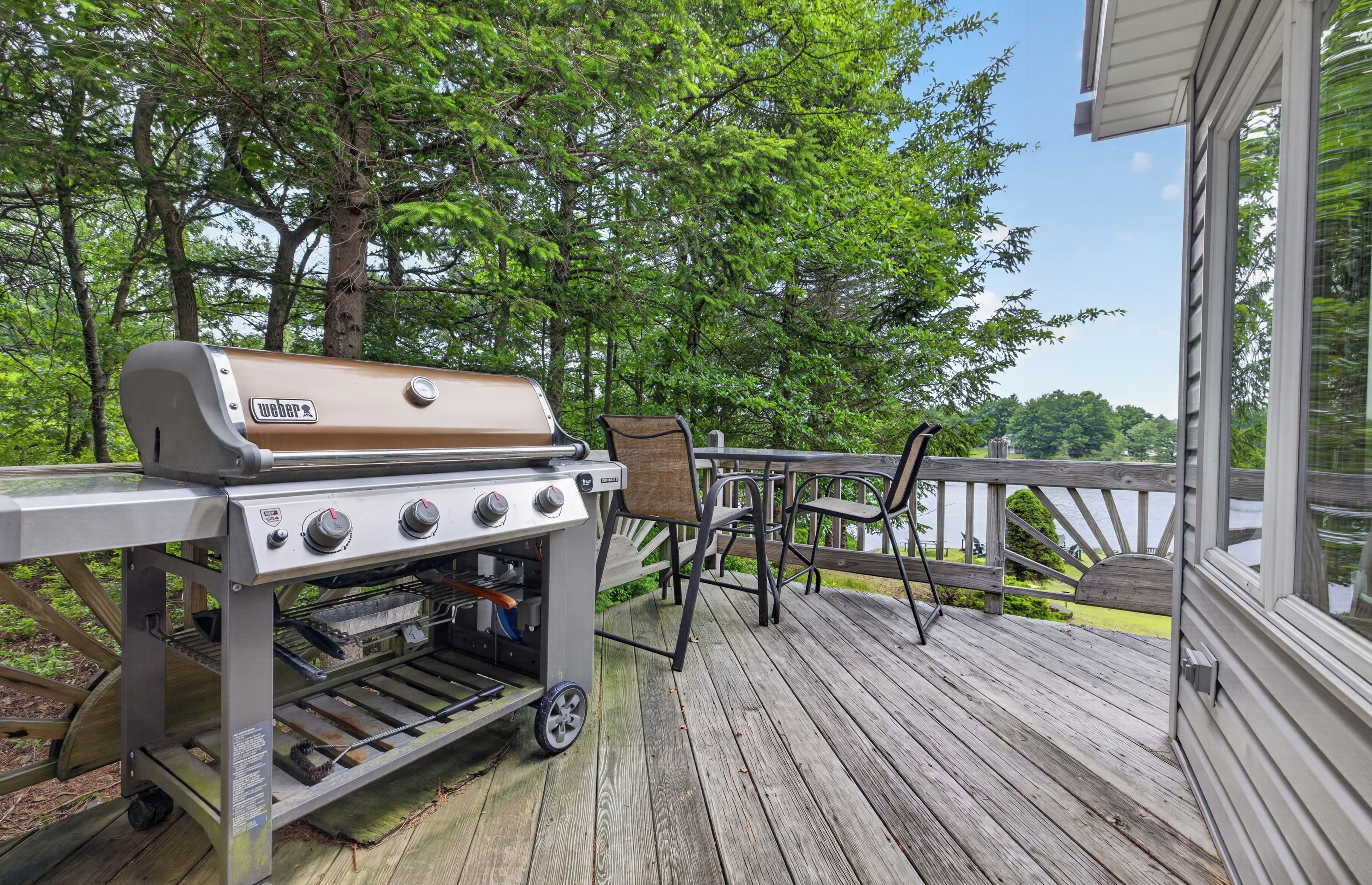 Una parrilla Weber sobre una terraza de madera, con vistas a árboles y una casa.
