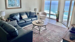 Una sala de estar con un sofá azul, una mesa de centro redonda, un ventilador de techo, un cuadro enmarcado y vistas al océano a través de amplios ventanales.