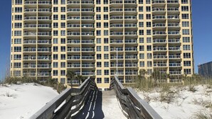 Un hotel de gran altura con múltiples balcones, una playa arenosa en primer plano y un cielo azul despejado.