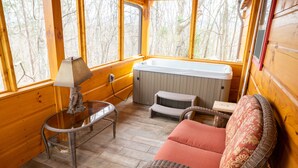 A cozy cabin interior with a wicker chair, a glass table with a lamp, a hot tub, and large windows offering a view of the forest. - NEW! Custom Cabin on 10 acres! (Sevierville)