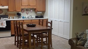 Un coin repas avec une table et des chaises en bois, une cuisine à l’arrière-plan et une fenêtre avec des stores.