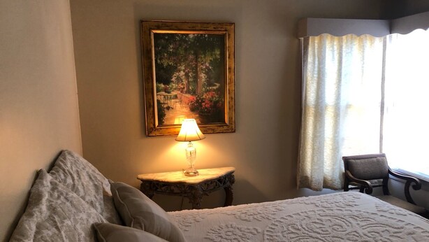 Una habitació d'hotel amb un llit, una làmpada, una cadira, un quadre a la paret i una finestra amb cortines.