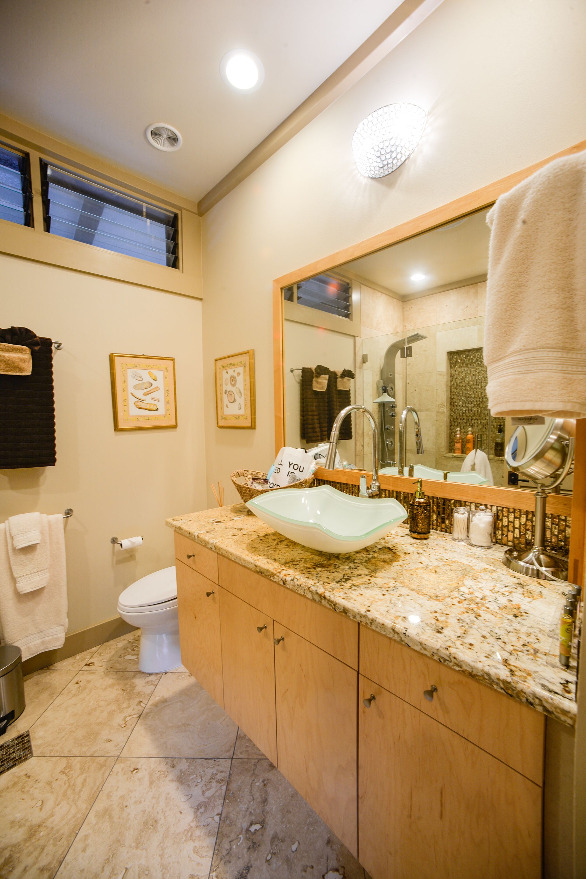 A bathroom with a large mirror, a sink, and a toilet. There are framed pictures on the wall and a towel hanging on the wall.