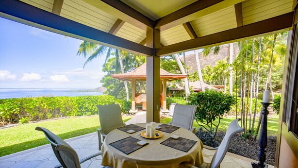 Une terrasse extérieure avec une table dressée pour deux personnes, offrant une vue sur un jardin luxuriant et l’océan.
