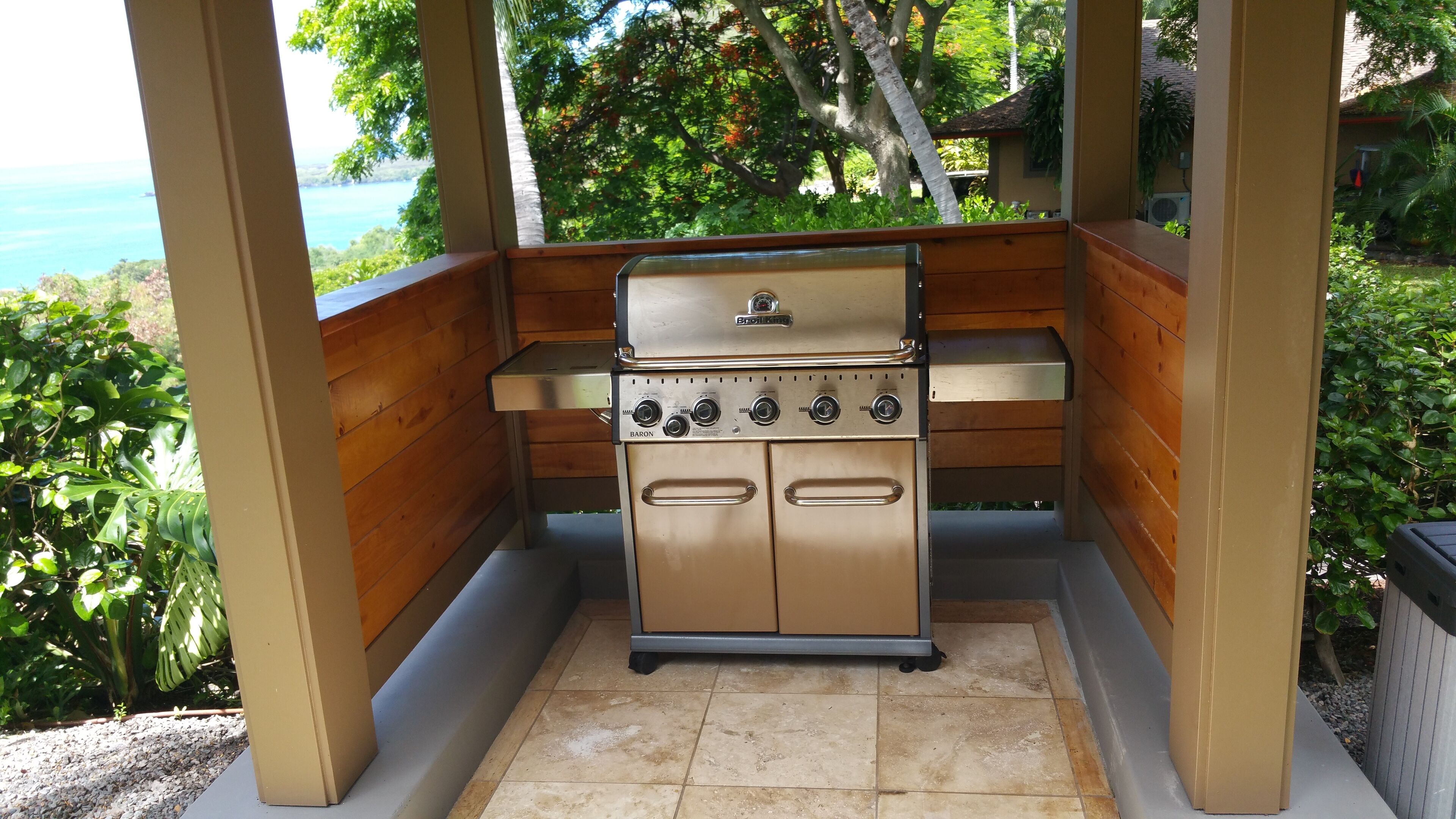 Una parrilla con cuatro quemadores y un panel de control, ubicada en una estructura de madera al aire libre con vista a un cuerpo de agua.