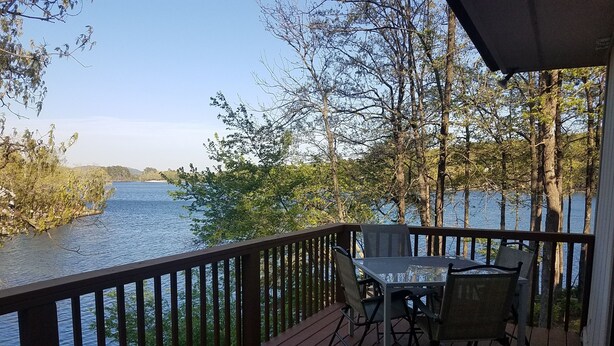 Een houten terras met een tafel en stoelen, met uitzicht op een rustige rivier, omringd door bomen.