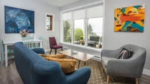 Uma sala de estar moderna com um sofá azul, uma poltrona cinza e uma mesa de jantar com cadeiras. Janelas amplas oferecem vista para o exterior.