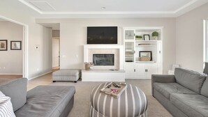 A modern living room with a flat-screen TV, grey sofas, a striped ottoman, and a white entertainment unit with shelves. - An Elegant Palm Desert Home, Great Nightly Rate! (Palm Desert)