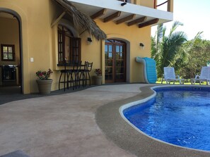 A pool area with a curved edge, outdoor seating with chairs, and a thatched roof structure. - Beautiful vacation home with ocean views nestled in the jungle with a pool (Chacala)