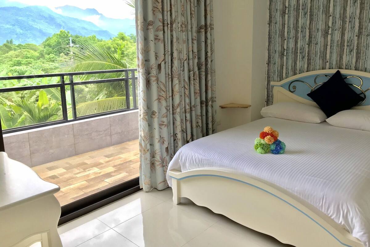 A bedroom with a bed, a nightstand, a window with curtains, and a view of greenery and mountains.