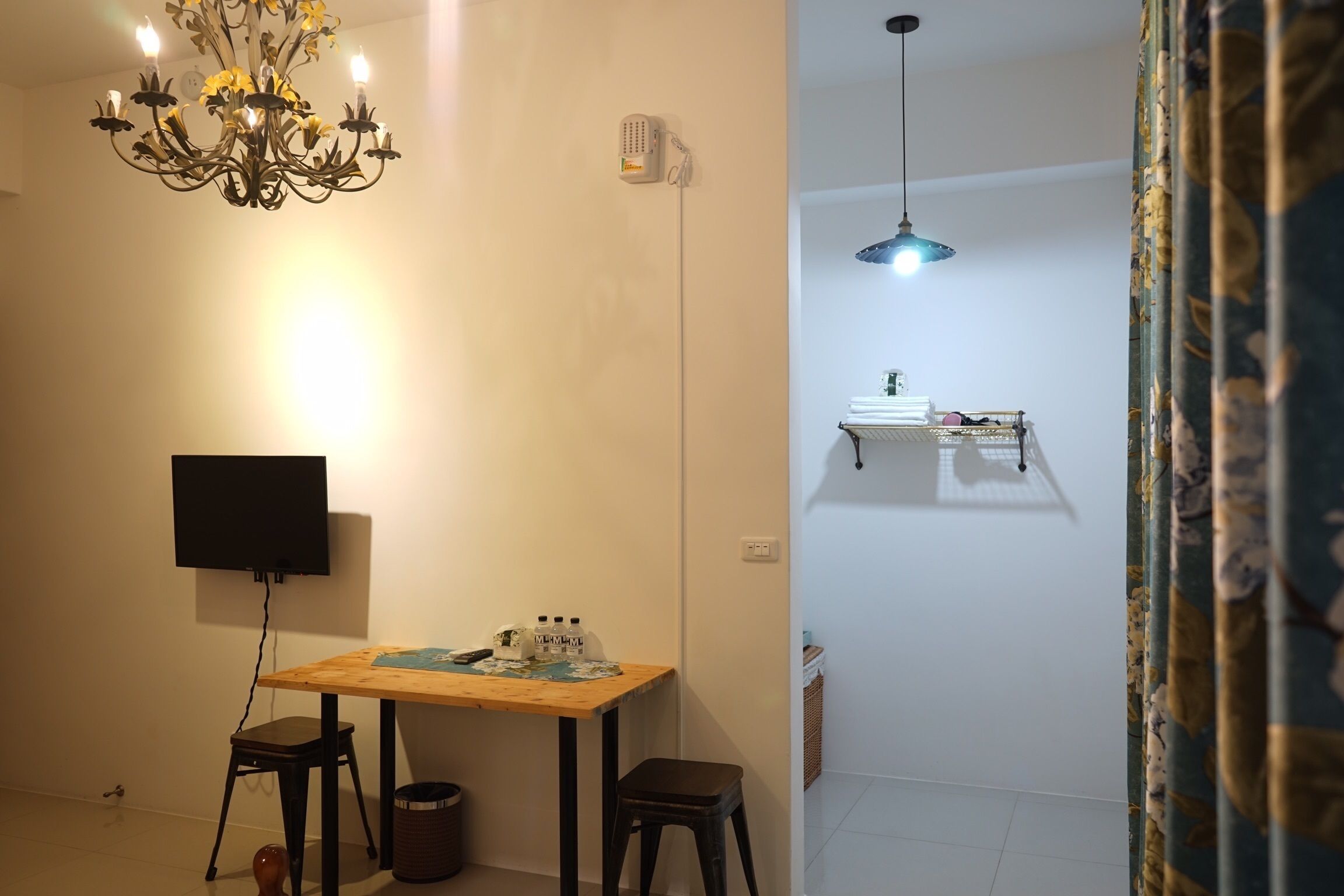 A room with a wooden table, two chairs, a TV, a chandelier, a ceiling fan, and a small shelf with items.