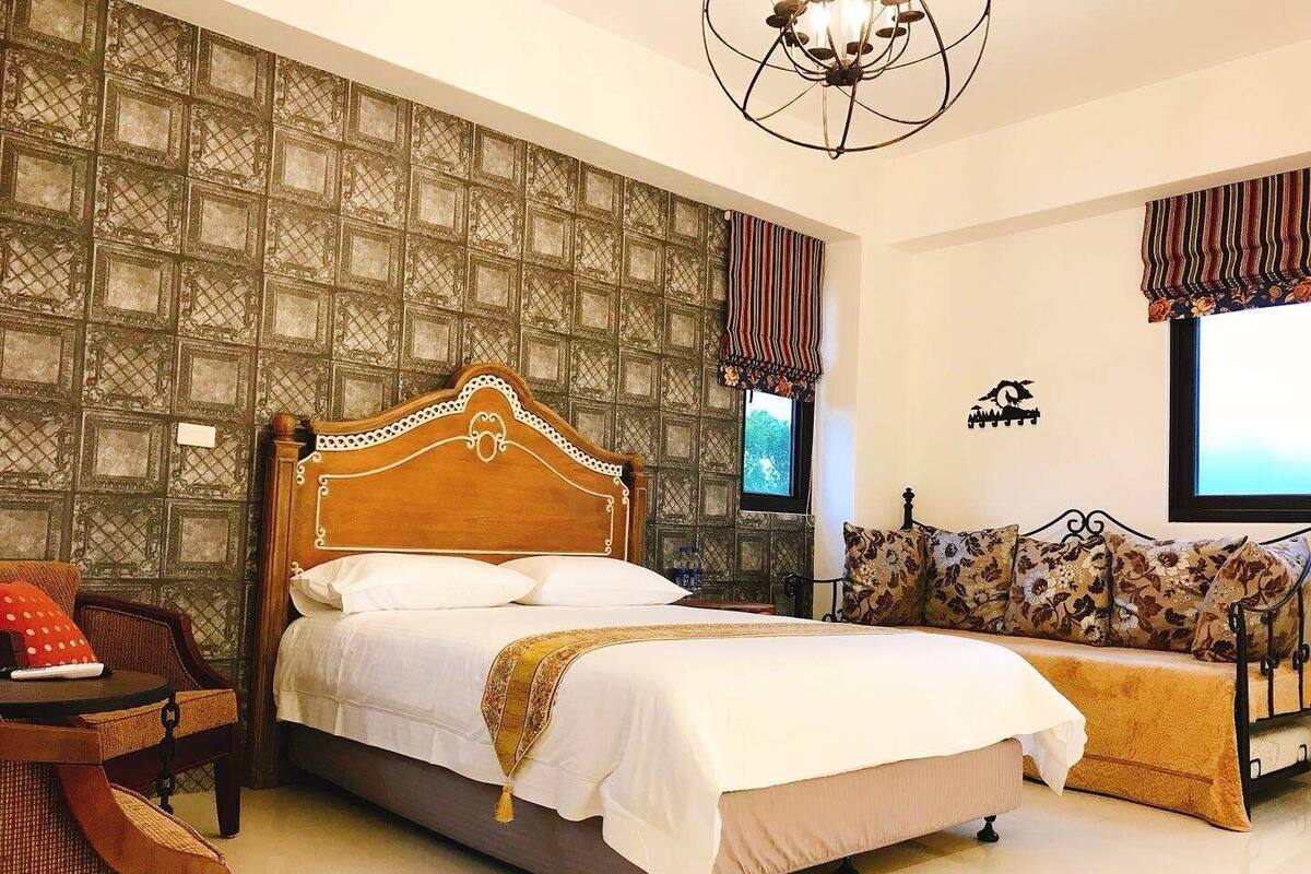 A bedroom with a large bed, a sofa, a chair, a chandelier, and a patterned wall.
