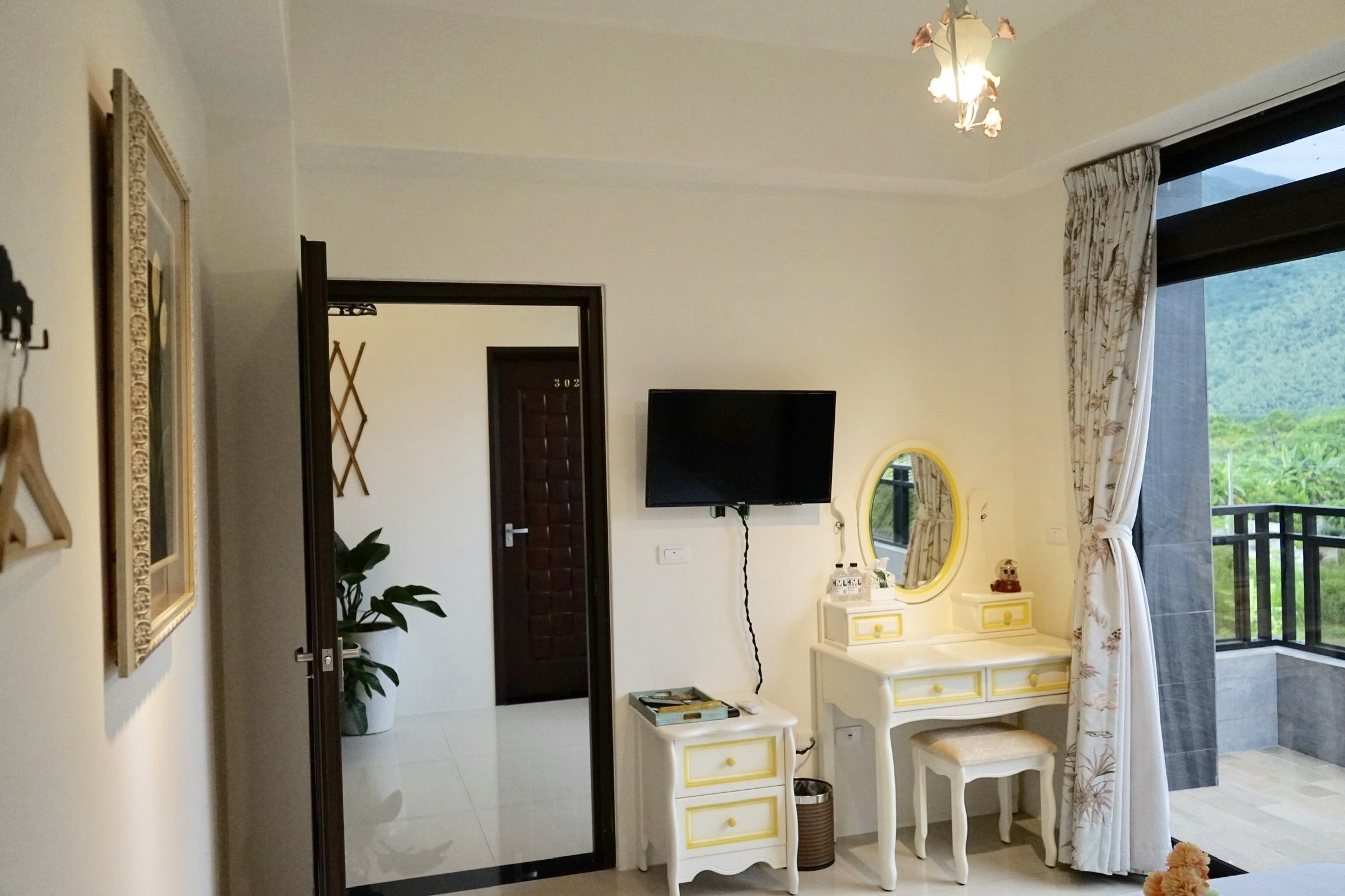 A bedroom with a white dresser, a mirror, a television mounted on the wall, and a window with curtains.