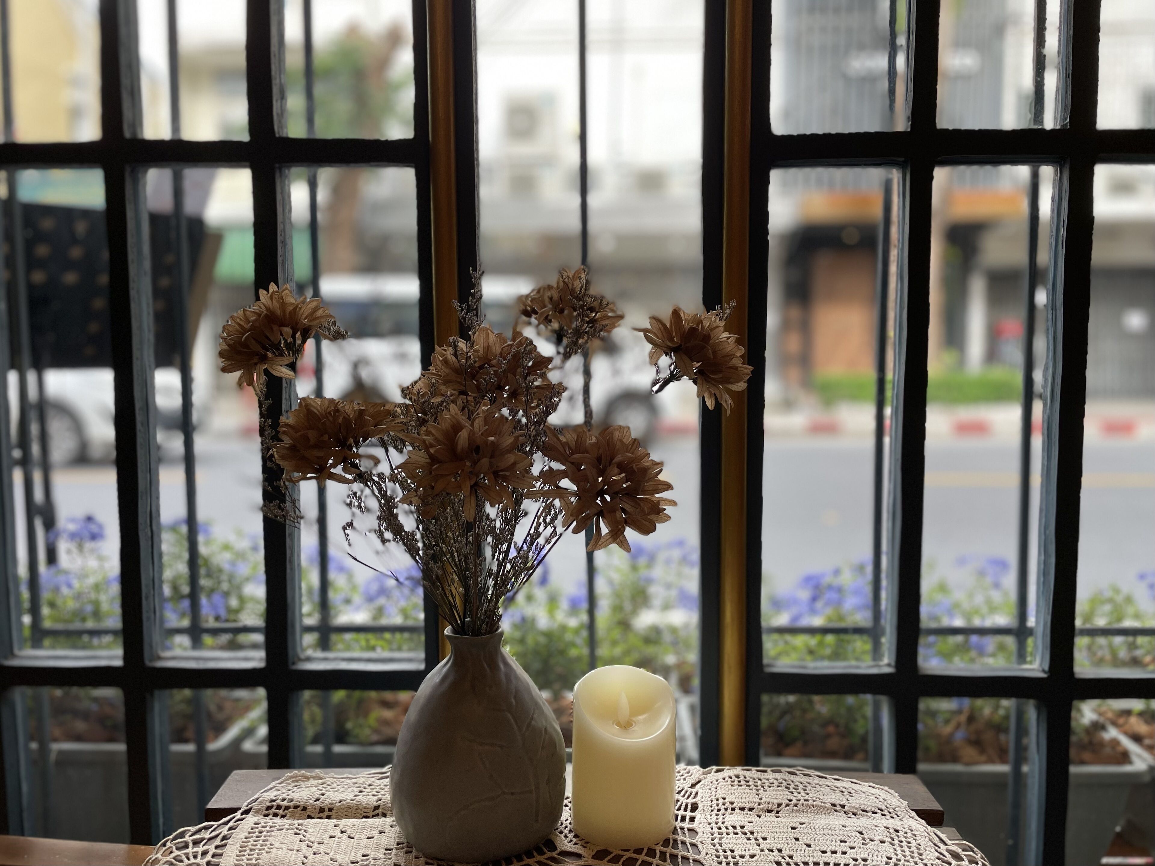 A vase with dried flowers and a candle on a table by a window with a view of the street.