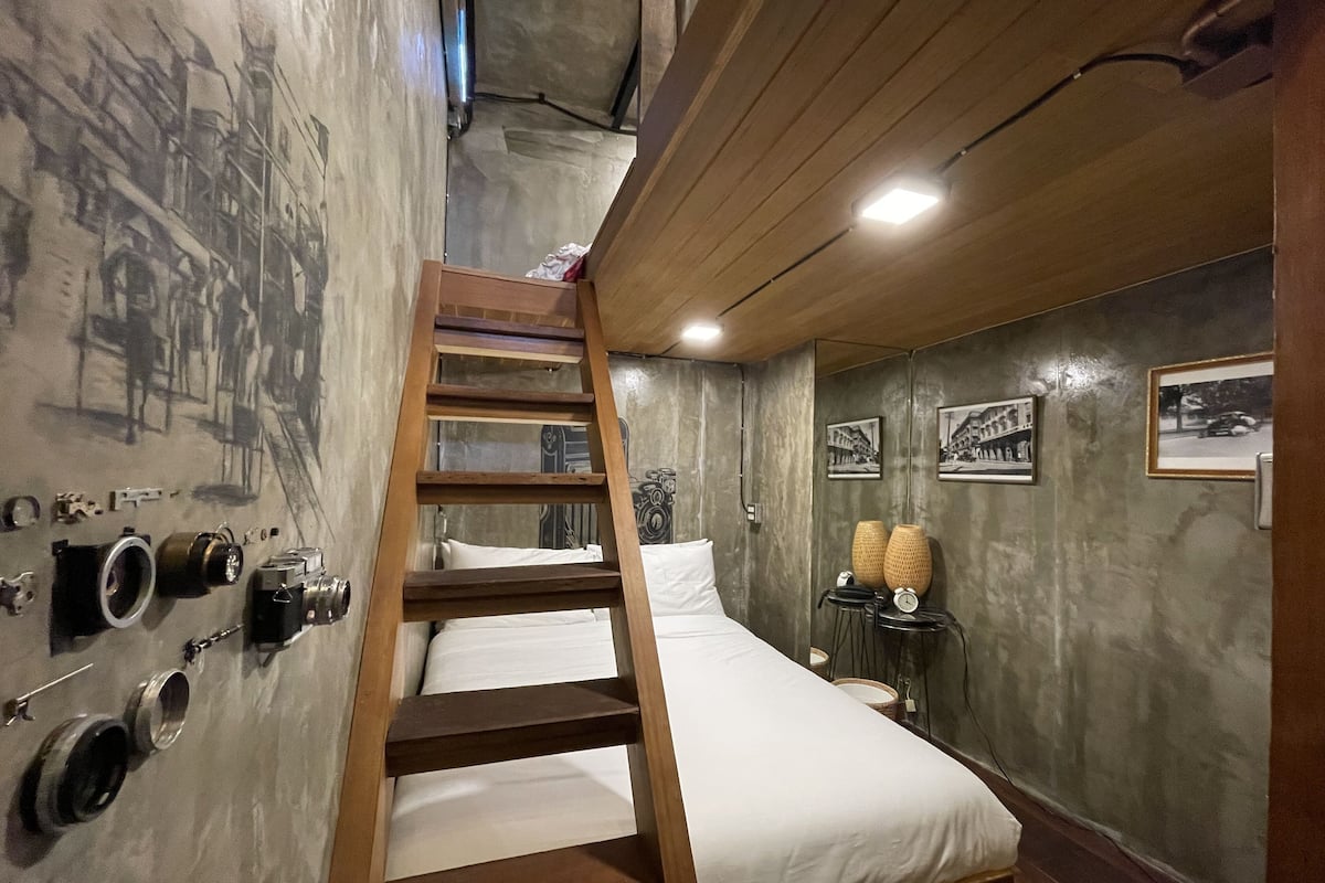 Een slaapkamer met een houten trap naar een stapelbed, een klein tafeltje met een klok, en oude foto's aan de muren.