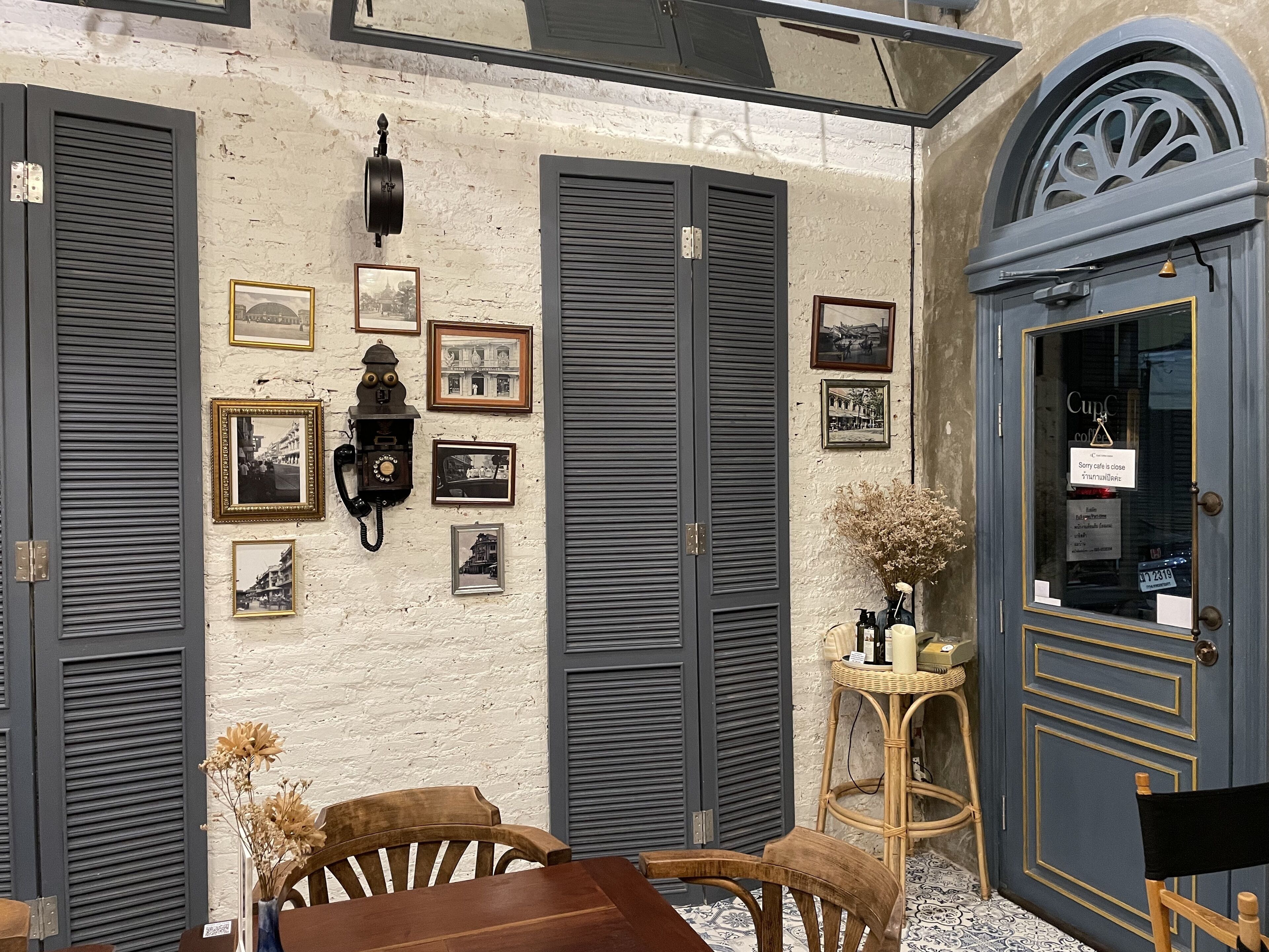 A café with a rustic interior, featuring a wooden table and chairs, a wall adorned with framed photographs, and a blue door with a sign indicating the café is closed.