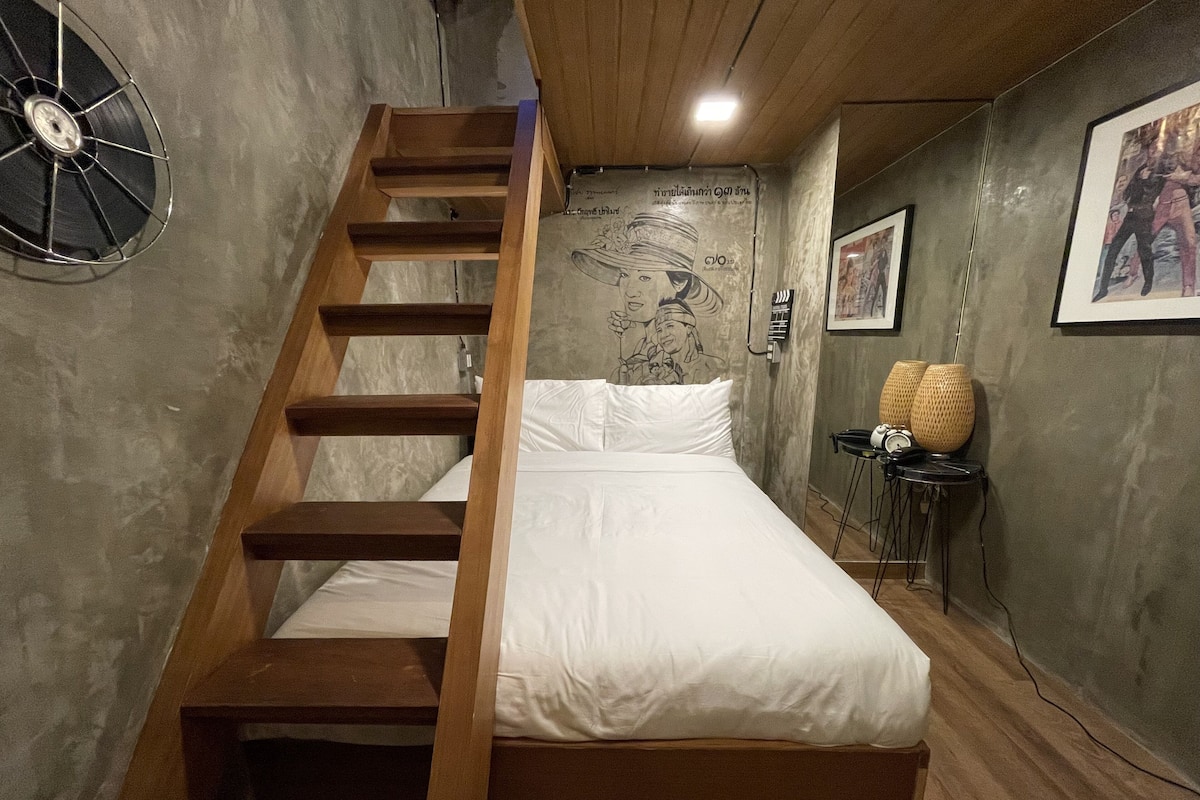 Een slaapkamer met een houten trap, een bed met wit beddengoed, een muurschildering en een kleine tafel met een lamp.