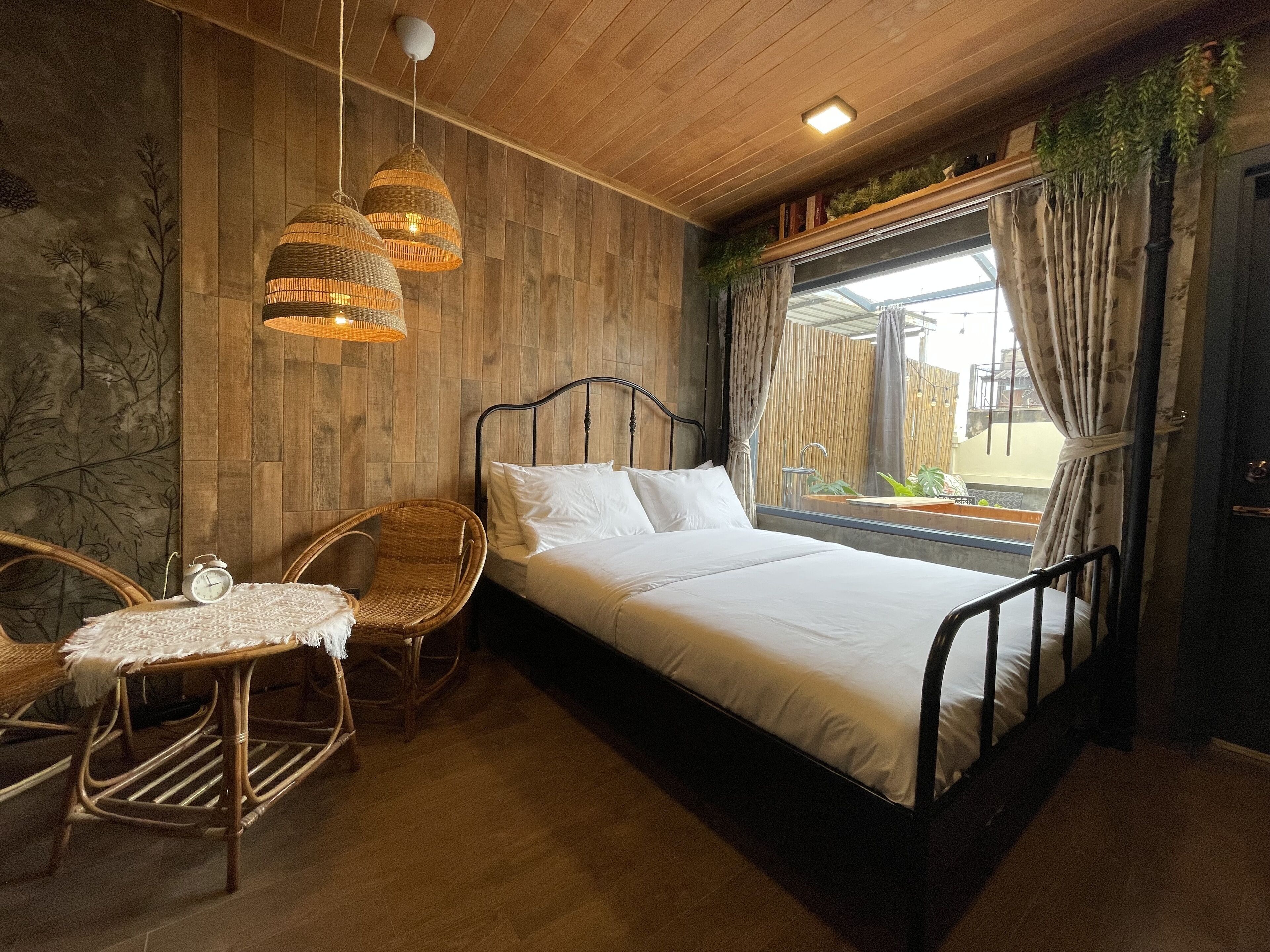 A bedroom with a bed, two wicker chairs, a small table, a wooden ceiling, and a window with curtains.