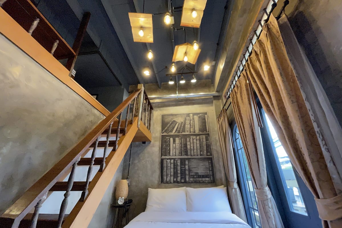 Een slaapkamer met een houten trap, een bed met witte lakens, een wandplank en een raam met gordijnen.