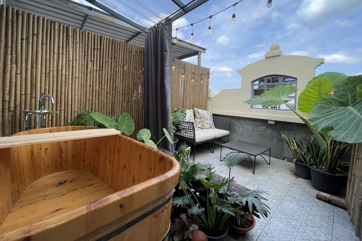 Een houten jacuzzi in een overdekte buitenruimte met bamboevloeren, potplanten en een zithoek met kussens.