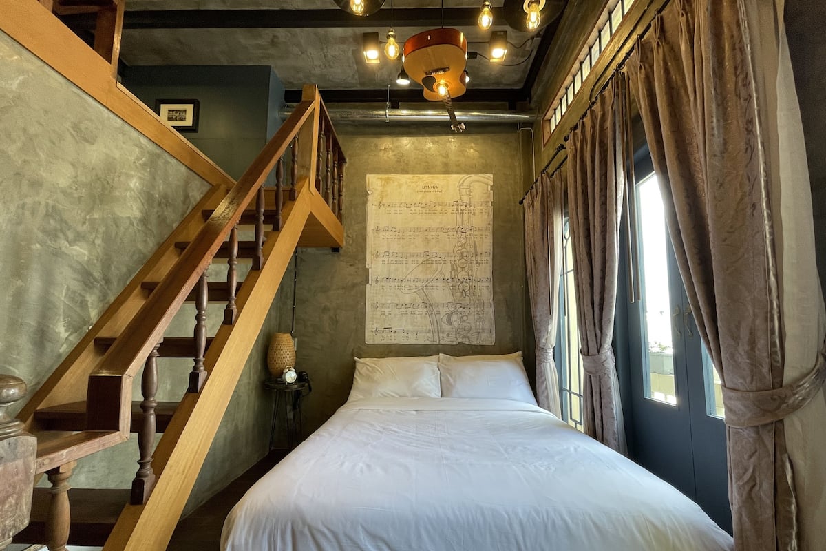 Een slaapkamer met een houten trap, een bed met witte lakens, een wand met bladmuziek en een raam met gordijnen.