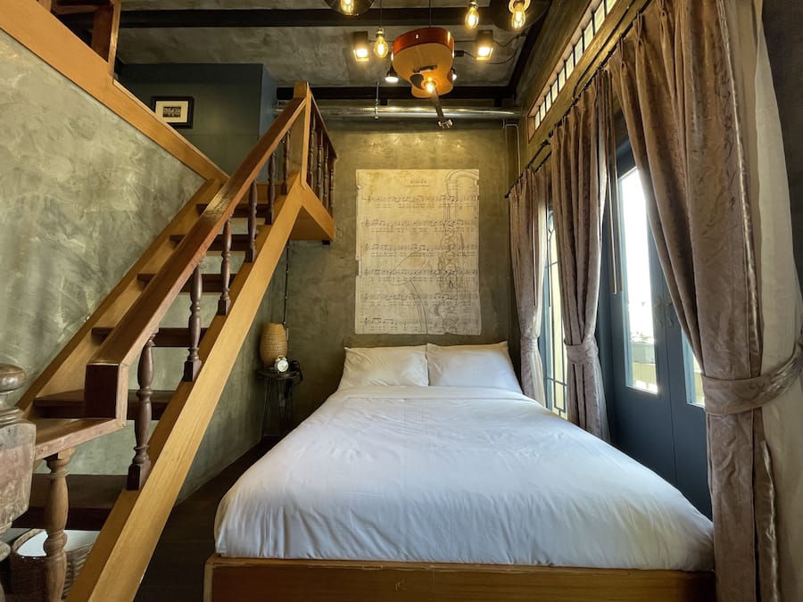 A bedroom with a wooden staircase, a bed with white linens, a wall with sheet music, and a window with curtains.