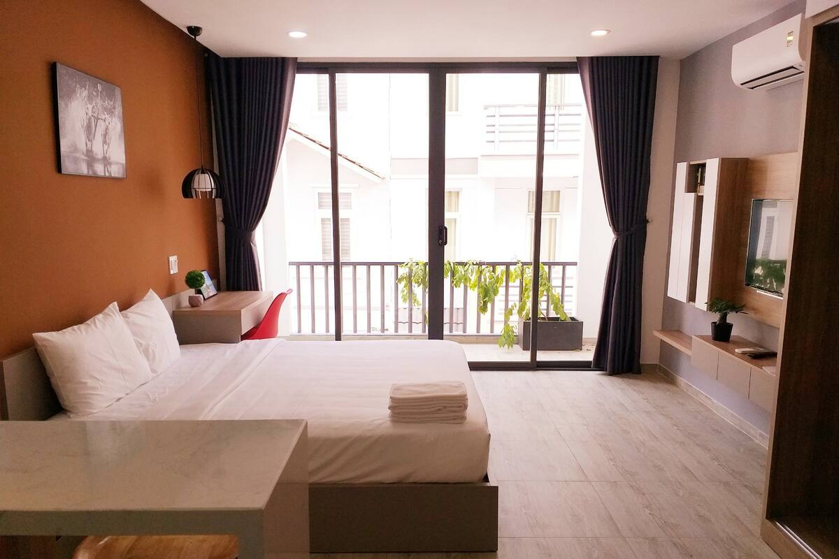 A modern hotel room with a bed, a desk, a chair, a TV, and a balcony with plants.