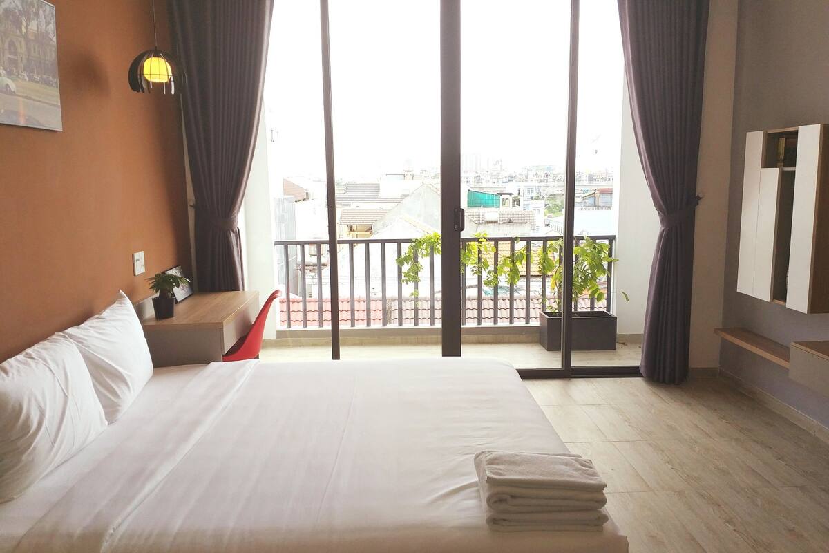 A hotel room with a large bed, a balcony with a view of the city, a small table with a plant, and a mounted TV.