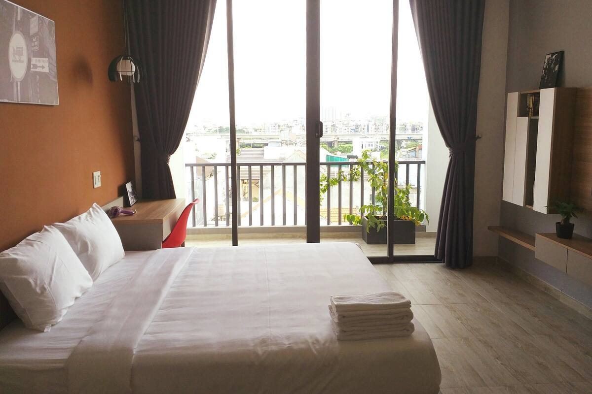 A hotel room with a large bed, a desk, a chair, a balcony with a view of the city, and a potted plant.