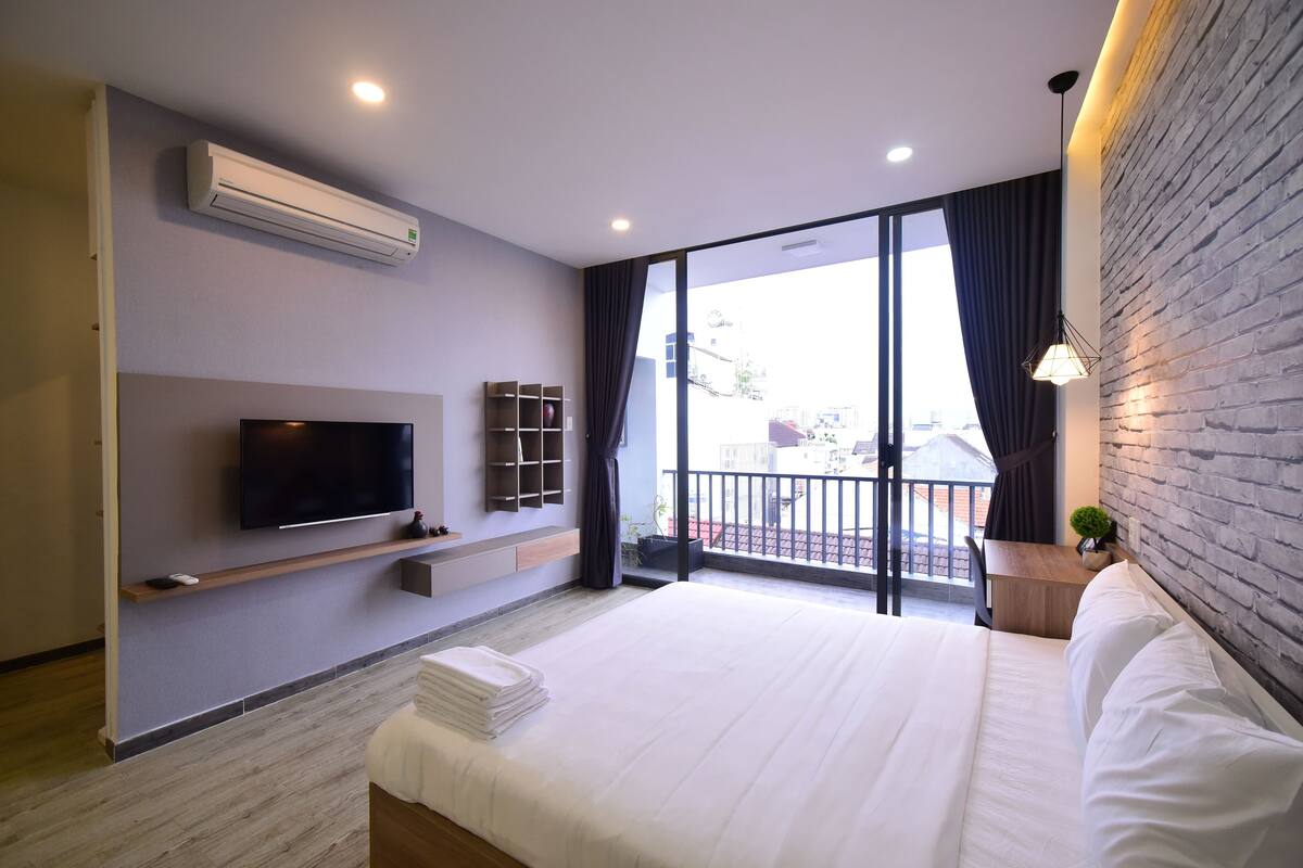 A modern hotel room with a large bed, a flat-screen TV mounted on the wall, a small seating area, and a balcony with a view.