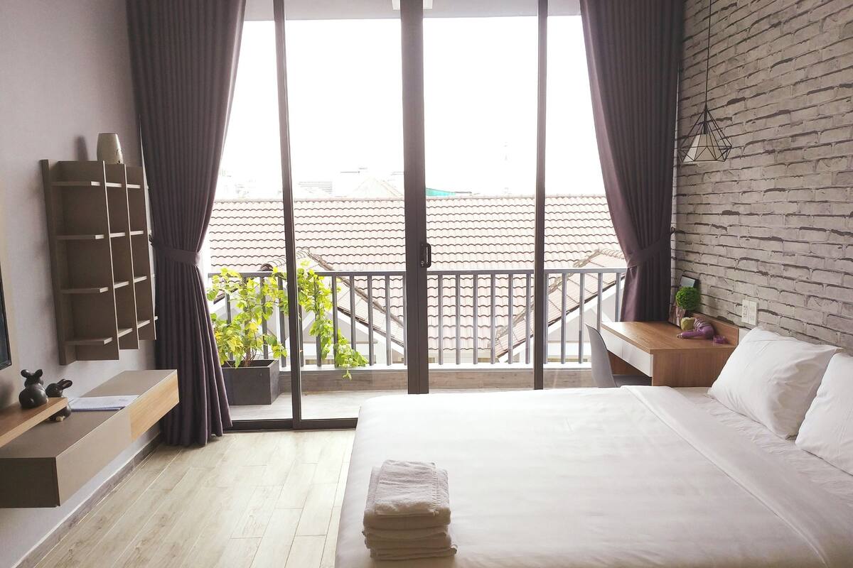 A hotel room with a large bed, a wooden nightstand, a small table, a wall-mounted shelf, and a balcony with a view of the city.