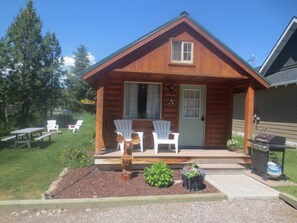 A wooden cabin with a porch, two Adirondack chairs, a welcome sign, and a small garden area. - A charming cabin close to Glacier National Park, Flathead Lake, and ski resorts. (Columbia Falls)
