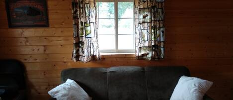 A wooden cabin interior with a sofa, two pillows, a framed welcome sign, and a window with patterned curtains.