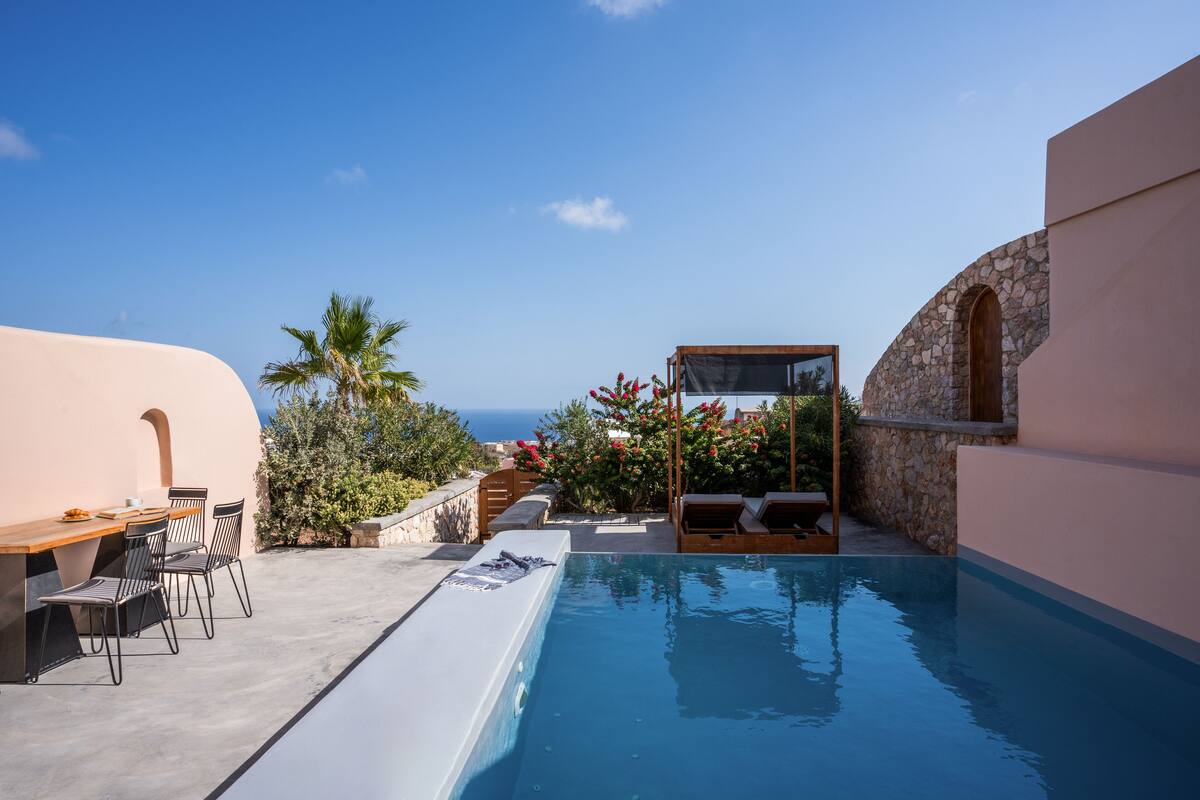 A pool area with a view of the sea, a wooden table with chairs, and a stone wall.
